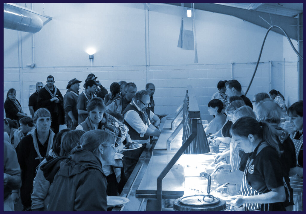The staff catering hall filled with lots of people and staff serving food.
